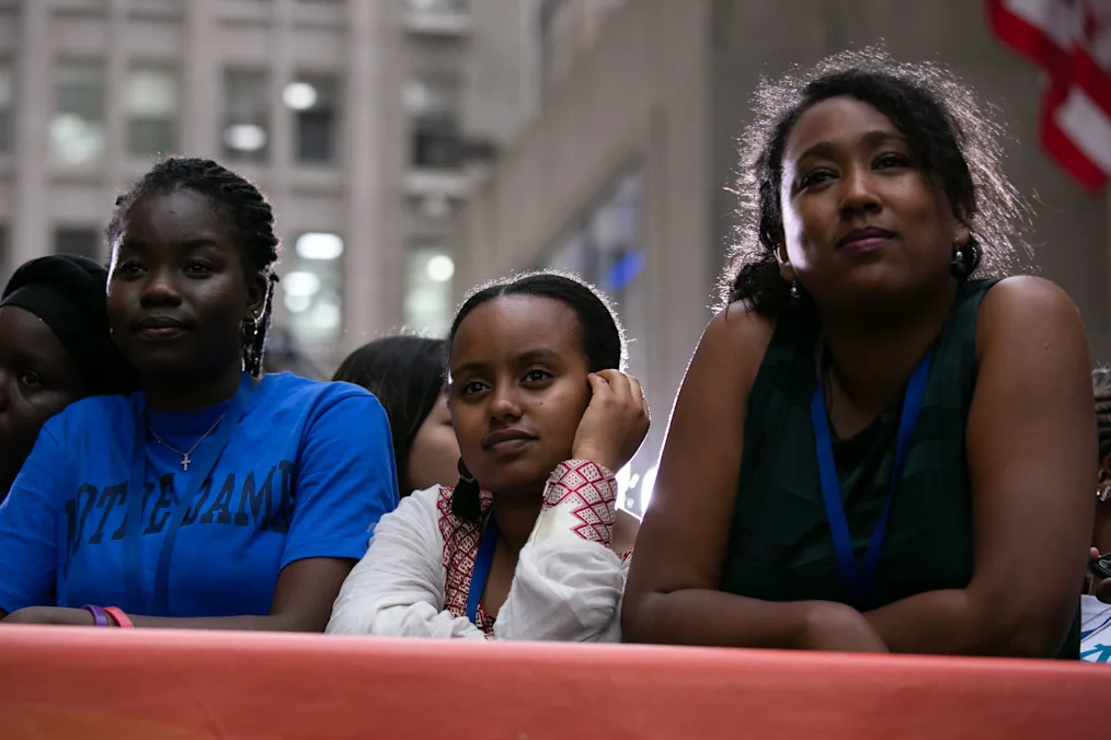 A small group of women with medium-deep to deep skin tones looking off to their right. They all have blue lanyards on. Out of focus in the background is a tall office building.