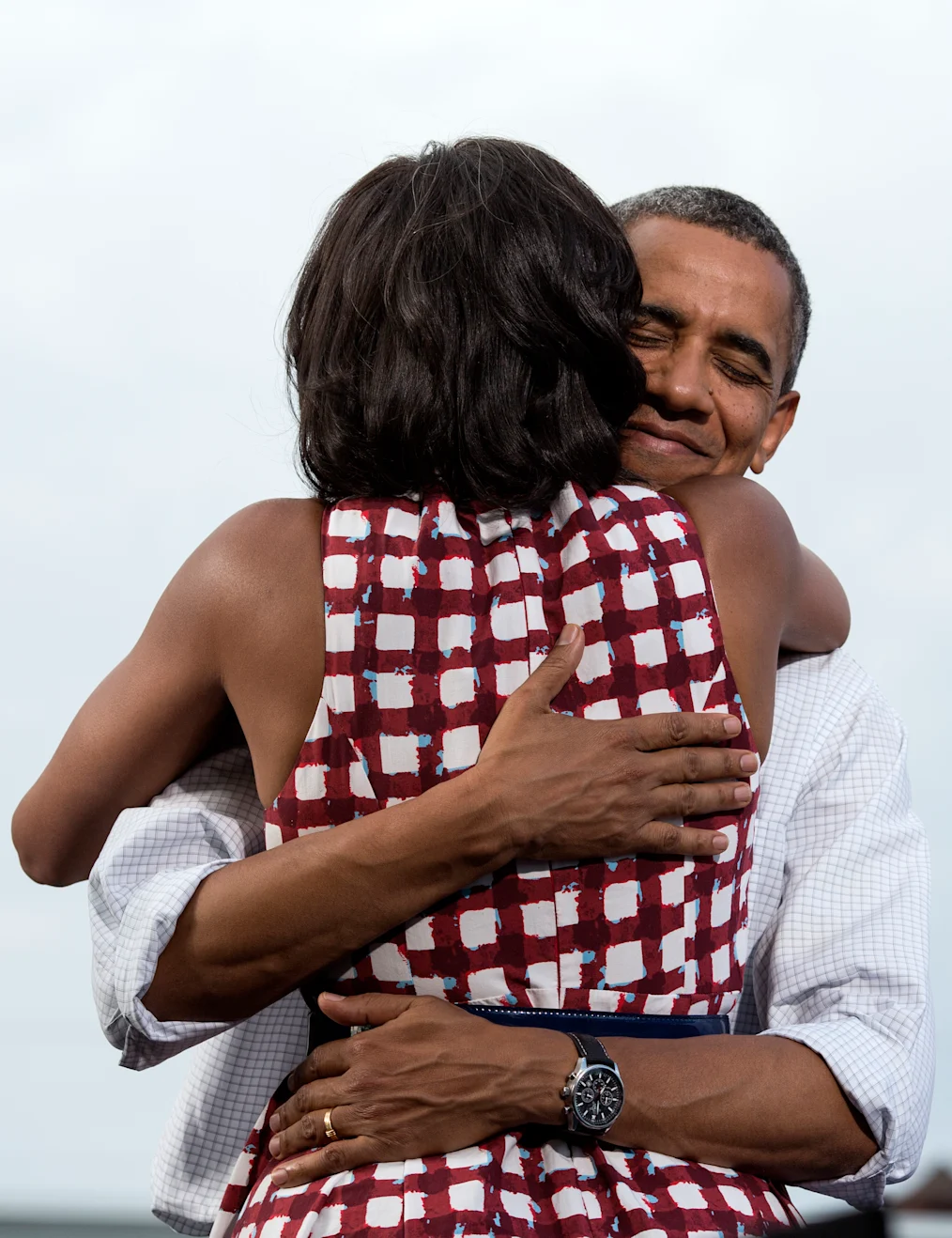President Obama holds a closed lip and eye smile as he hugs his wife. Mrs. Obama isn’t facing the camera. She is wearing a red and white patterned dress.