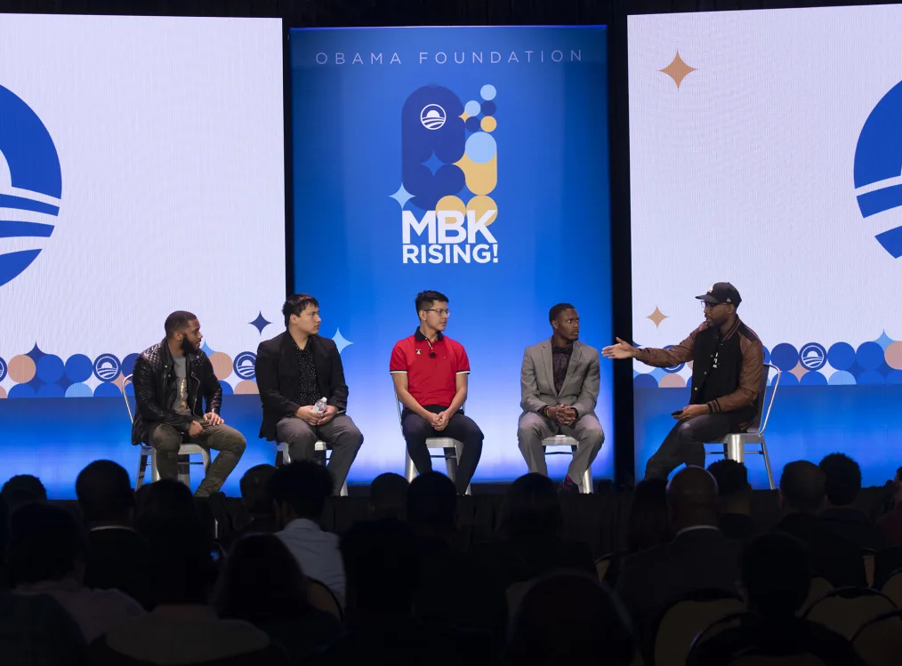 Five men sit on metal chairs on a stage. They are all looking to the right where one appears to be motioning with his hands. Behind them is a large blue, white and yellow sign with the words "Obama Foundation, MBK Rising."