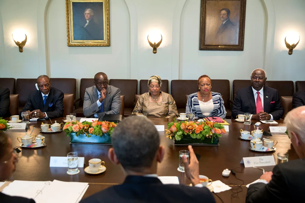 President Obama sits across five individuals with different skin tones at a long brown table with two pink flower bouquets in the center of the table. There is a white wall in the background with two portrait paintings and three lamps.