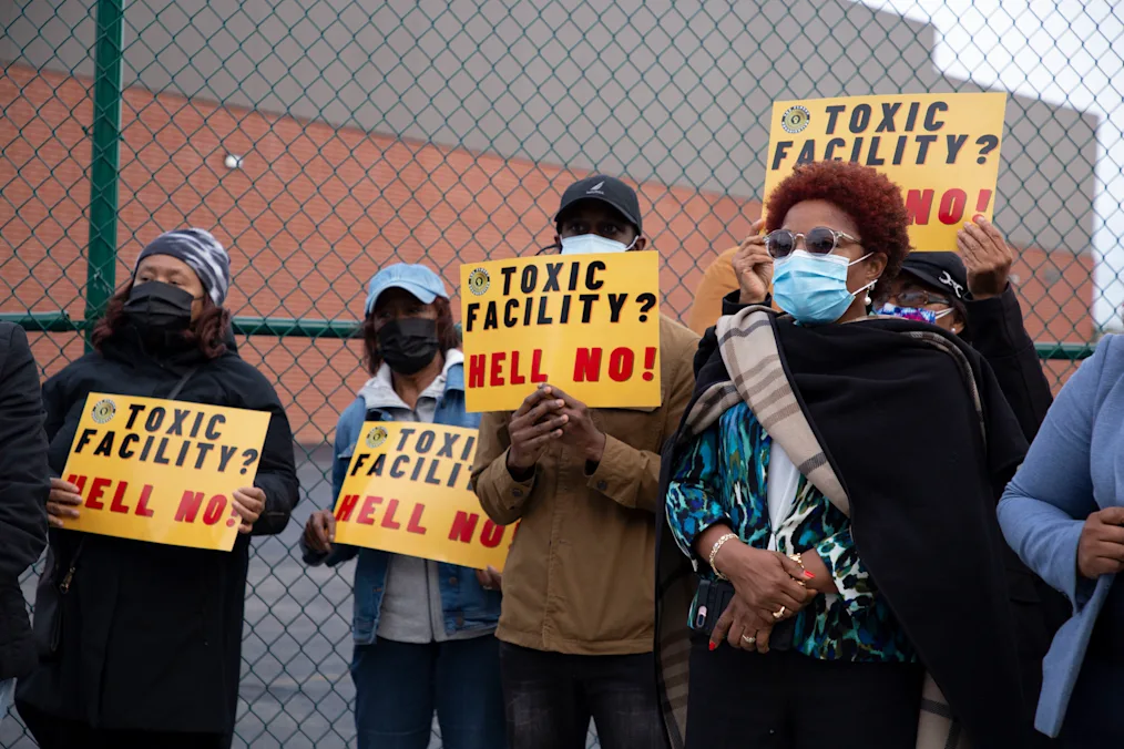 A group of individuals, with a range of light to dark skin tones, hold yellow posters that read,  “Toxic Facility? Hell No!”