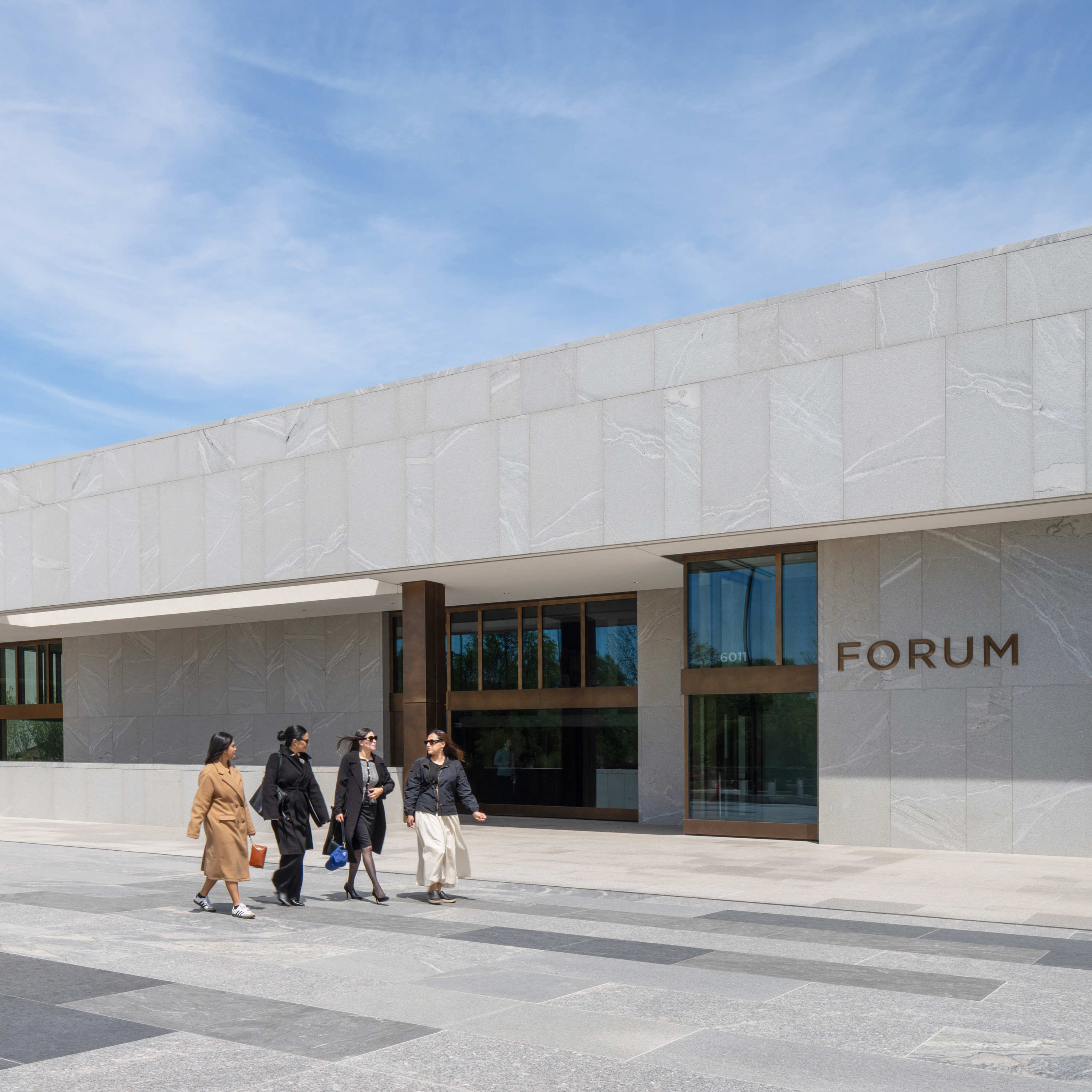 Four women walk together on a stone plaza past the entrance of a modern granite building labeled "FORUM." On the far right, bronze-toned, three-dimensional capital letters mounted on the wall spell out "FORUM". Beneath the sign, floor-to-ceiling glass windows and doors framed in dark bronze form the main entrance