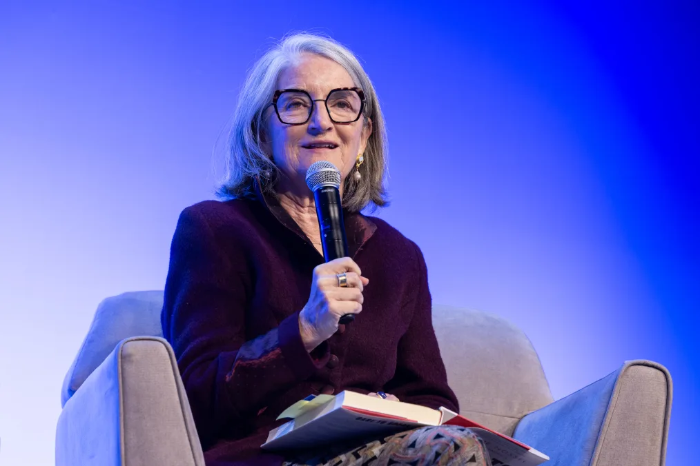 Ellen Alberding, a woman with a light skin tone and short gray hair, holds a microphone to her mouth as she sits on a stage. The background is blue. She is wearing a burgundy jacket and glasses. 