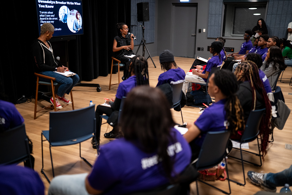 In a dark gray media room, a Black woman sits on a chair in front of a screen and talks into a microphone. Another Black woman sits across from her on the left. An audience of Black youth in purple shirts sit in rows facing the speaker and take notes on clipboards.