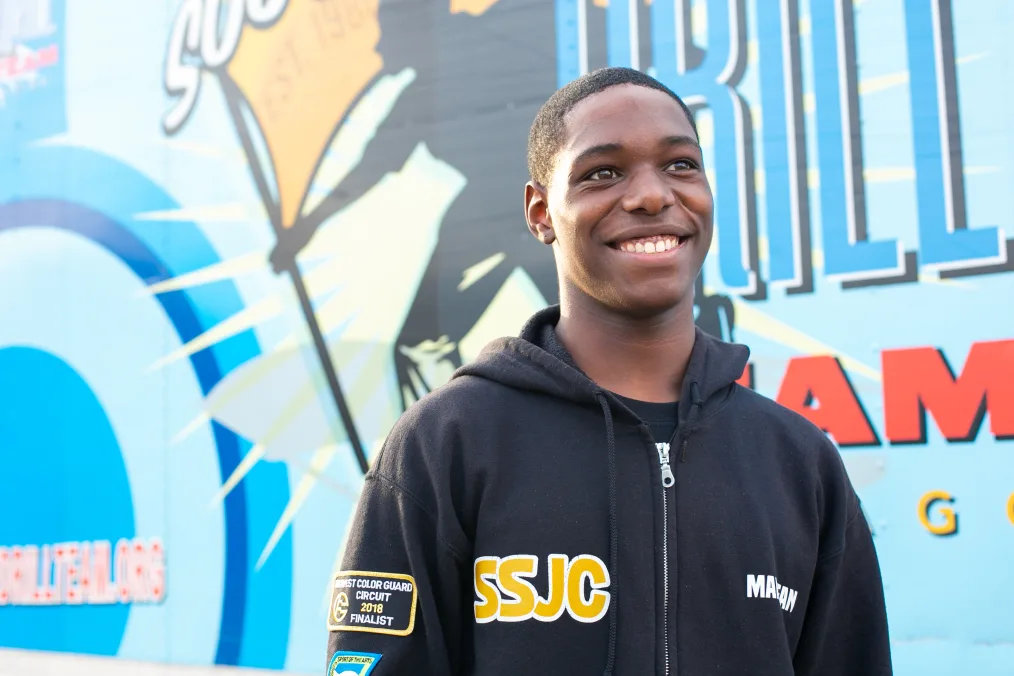 A young Black man looks slightly to the right, smiling widely. He wears a zip-up hoodie and stands in front of a colorful wall in unintelligible graphics.