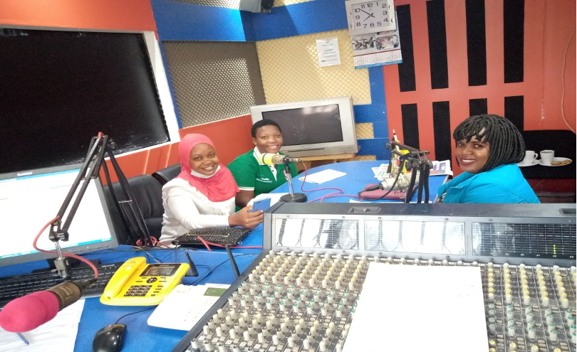 A radio host in a pink hijab smiles, Susan Sabano smiles, and another radio host with neck length individual braids smiles at the camera. All women are of deep skin tones and are sitting in a studio surrounded by microphones and radio production equipment.