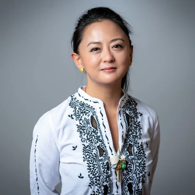 A woman with black hair pulled back looks at the camera. She wears gold earrings and an intricately embroidered black-and-white top. Around her neck is a necklace displaying several colorful objects.