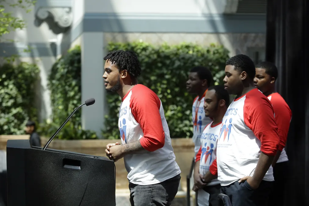 

A man with a deep medium skin tone with short black twists stands in front of a podium wearing a red and white shirt while other men ranging in medium to deep skin tones stands in the background 