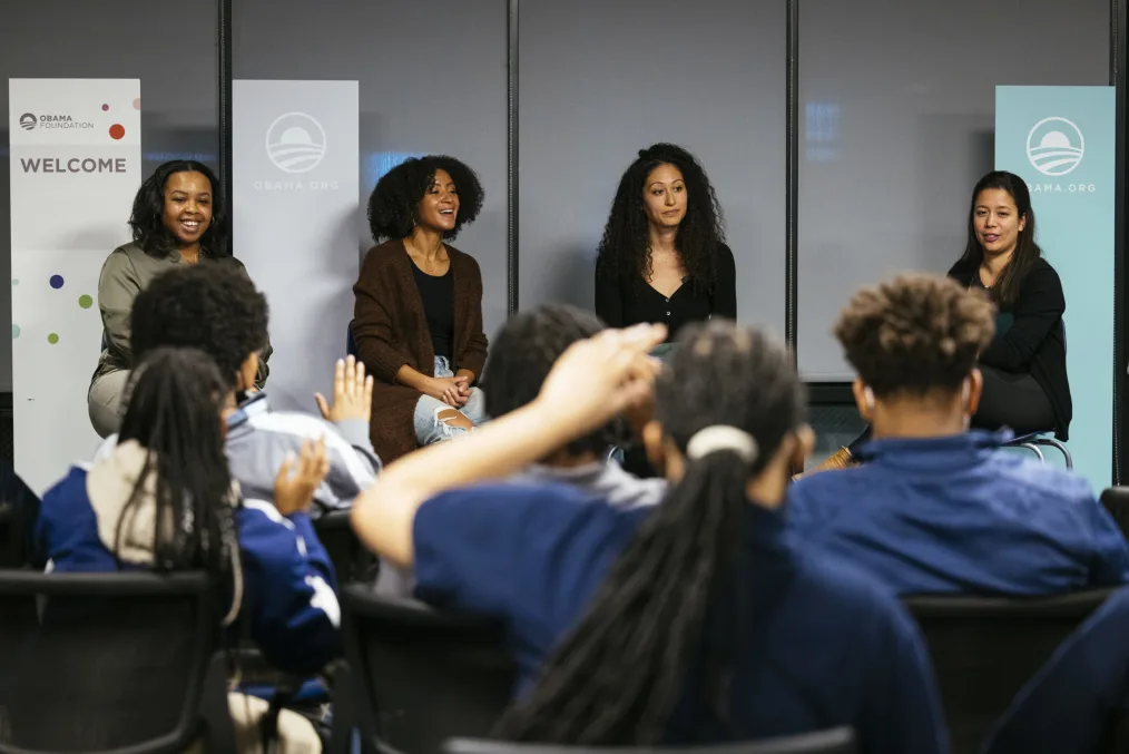 Four women, with medium to medium-light skin tone and business casual attire, talk with a group of students during an Obama Foundation event.