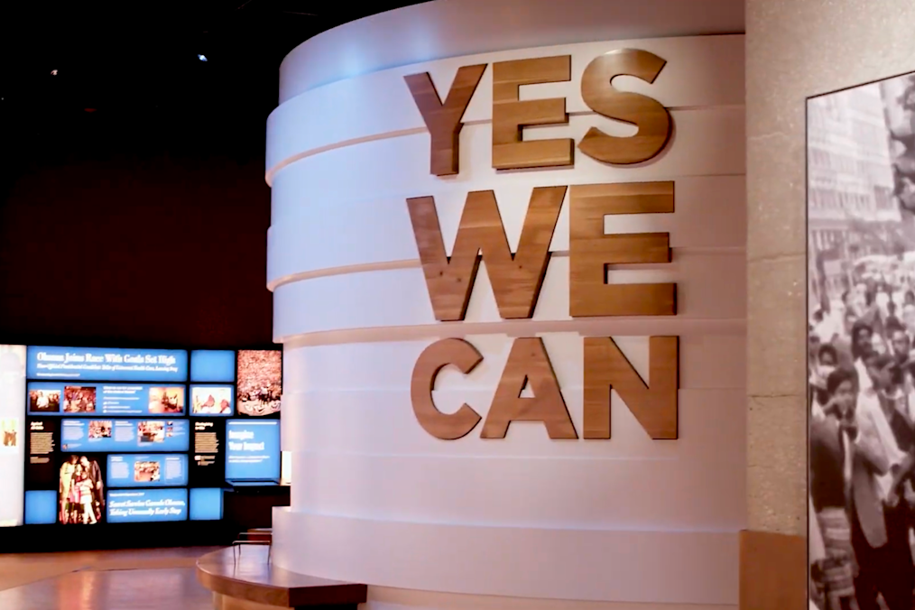 An image of the “Yes We Can” exhibit inside the Museum Building. The exhibit is a large circular structure with words that say “Yes We Can” in wooden letters. An exhibit screen is in the background.  