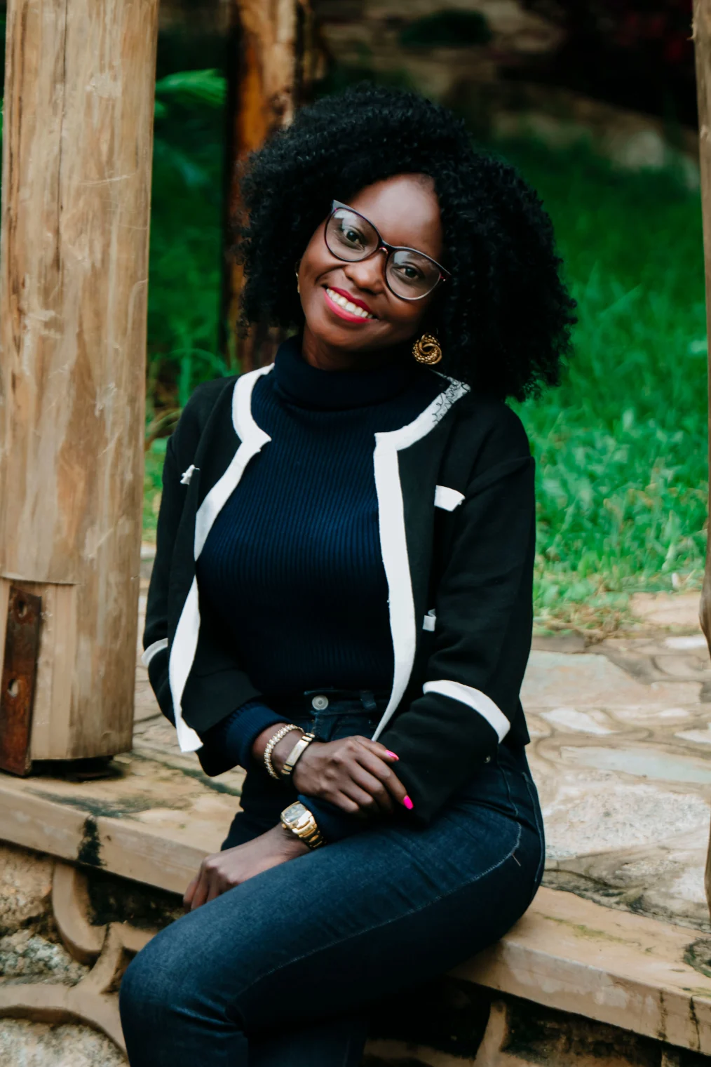 A Black woman with a deep skin tone sits on a wooden step outdoors. She has curly black hair and is wearing glasses as she smiles at the camera. The woman is wearing a black sweater with white lining, a black turtleneck, and denim jeans. Her fingernails are painted bright pink and on her left wrist is a watch and on her right wrist are two gold bracelets.