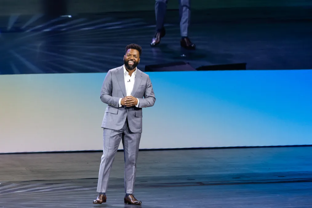 Democracy Forum host Baratunde Thurston, a Black man with a deep skin tone, stands on stage at the 2023 Obama Foundation Democracy Forum. He is wearing a gray suit. 