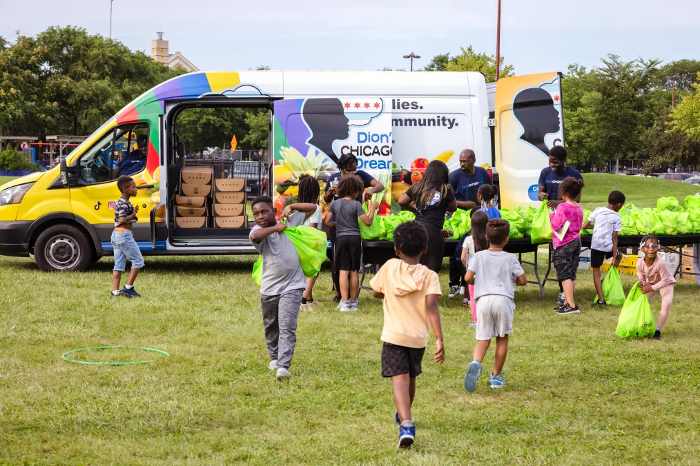 Children with a range of light to deep skin tones gather around a wrapped van. The children are holding green plastic bags filled with goodies. The truck in the background reads, “Dion’s Chicago Dream.” 