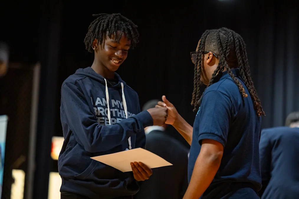 A boy with dark skin tone smiles as he holds a certificate and shakes hands with an almond-skin toned man.