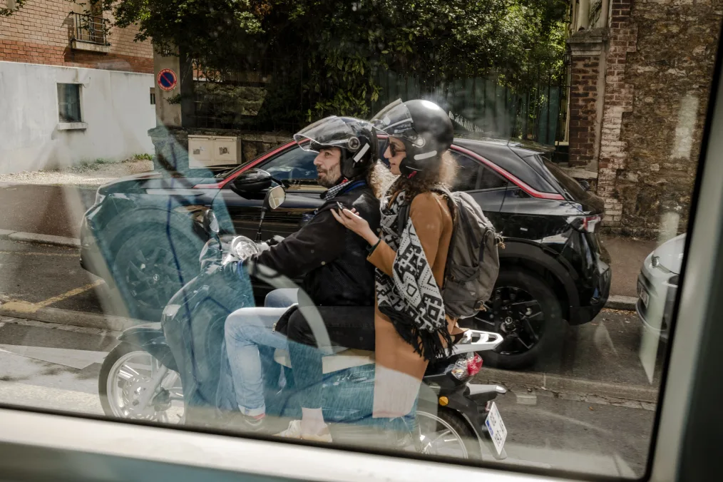 A man with a light skin tone wearing denim jeans, a black leather jacket, and a black helmet is driving a black motorcycle with a woman with a light skin tone wearing a tan jacket, black jacket, and lack helmet sitting on the back of the motorcycle. In the background are a black car and a brick home. 