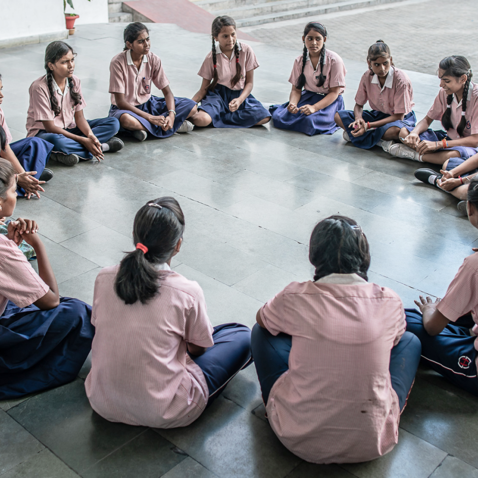 A group of young girls sit in a circle on the floor outside. Each of the girls is wearing a pink short sleeved blouse, a long dark blue skirt, and white sneakers. 