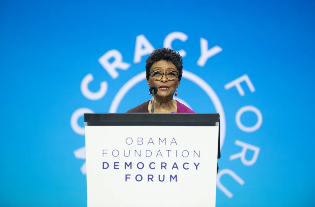 An older lady with medium-deep skin, short black hair, makeup, big camoflouge glasses, and dangling earings stands at a Obama Foundation Democracy Forum podium. She is giving a speech. She stands infront of a sky blue wall with the obama foundation logo blurred in the background.