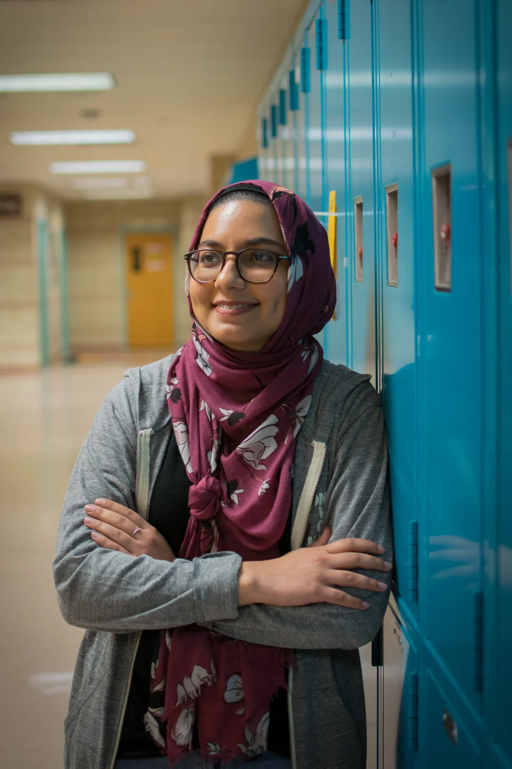 A medium skin toned Brown woman is leaning to the right against a set of blue lockers in a hallway. She is smiling with her teeth showing and looking away from the camera with her arms folded. She is wearing a multi-colored scarf that covers her hair and neck, a black T-shirt and a gray zippered hoodie.