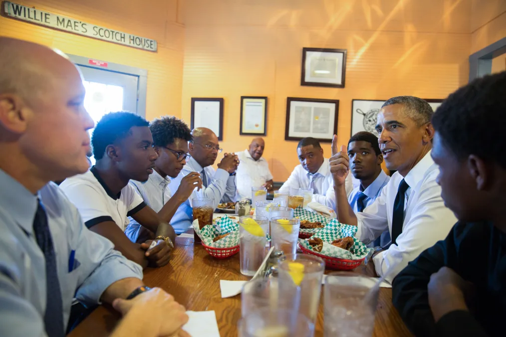 President Obama sits around a table with food and MBK mentees. All are dressed professionally and have a range of light to deep skin tones. 