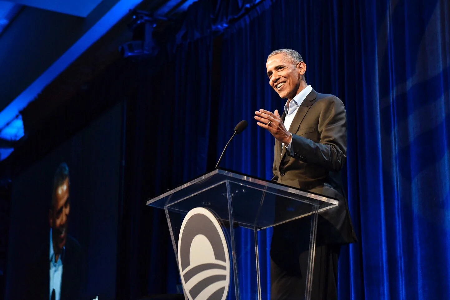 President Obama speaks on a microphone that is propped up on a clear glass stand with the Obama Foundation rising sun logo on the front.