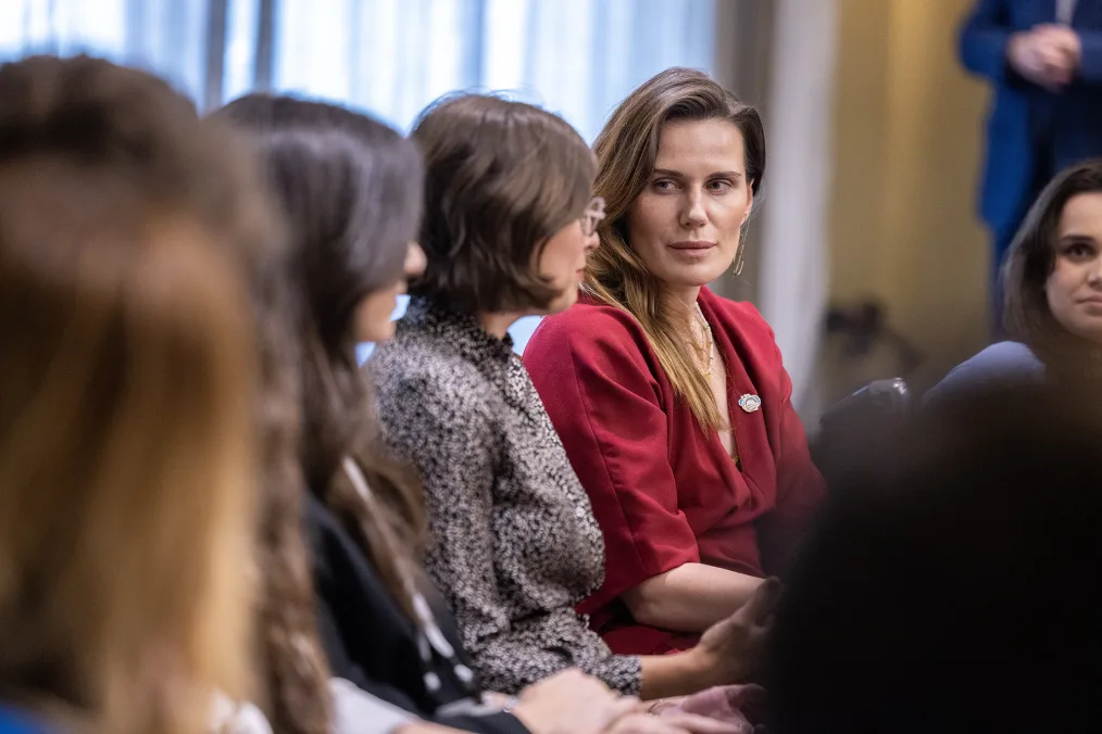 Women with light skin tones and a range of short and long brown hair sit at a table and talk. All are dressed professionally. 