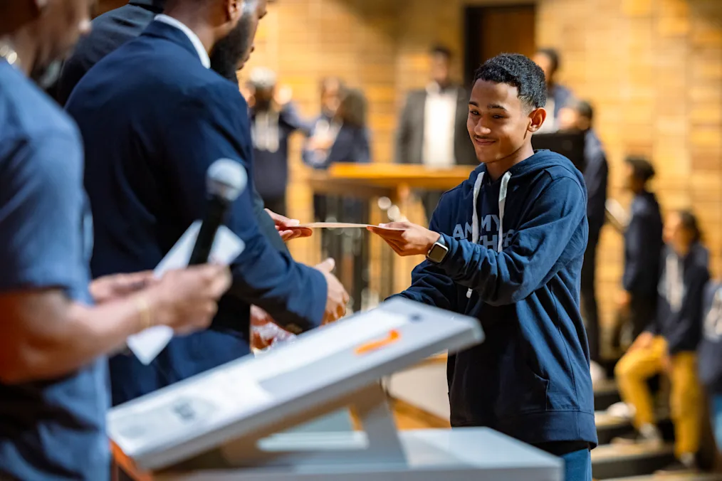 A boy with a medium skin tone grabs a certificate from a man with a medium skin tone. More young men with a range of light to dark skin tones are lined and blurred in the background.