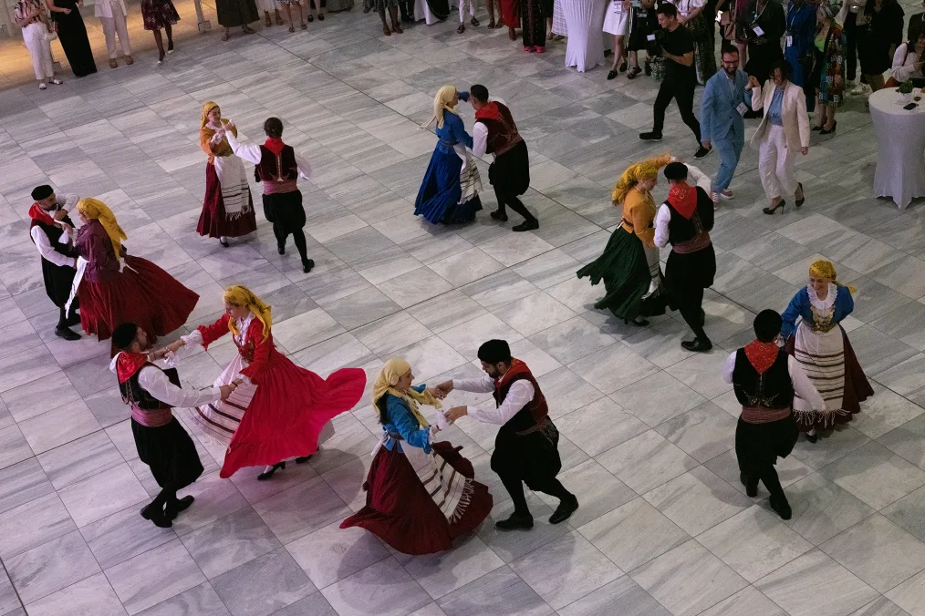 A troupe of traditional Greek dancers of varying skin tones, hold hands while they dance in groups of two. In the background there are people standing and watching.