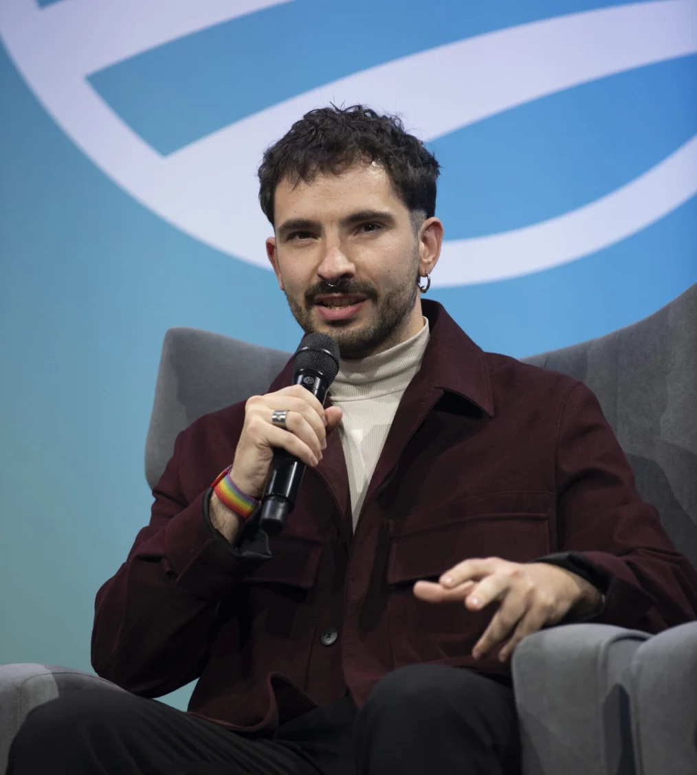 A man with light skin with warm undertones sits in a comfy grey chair in front of a huge sunset gradient of the Obama Foundation logo. He has tossled black wavy hair, a taper cut, a standard beard and mustache, and nose and ear circular piercings. He is wearing a bugandy collared jacket, with a beige turtle neck. He is holding a mic and saying something.  