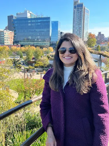 A close up photo of a woman with a neutral skin tone wearing shades in a purple jacket with a park and buildings behind her