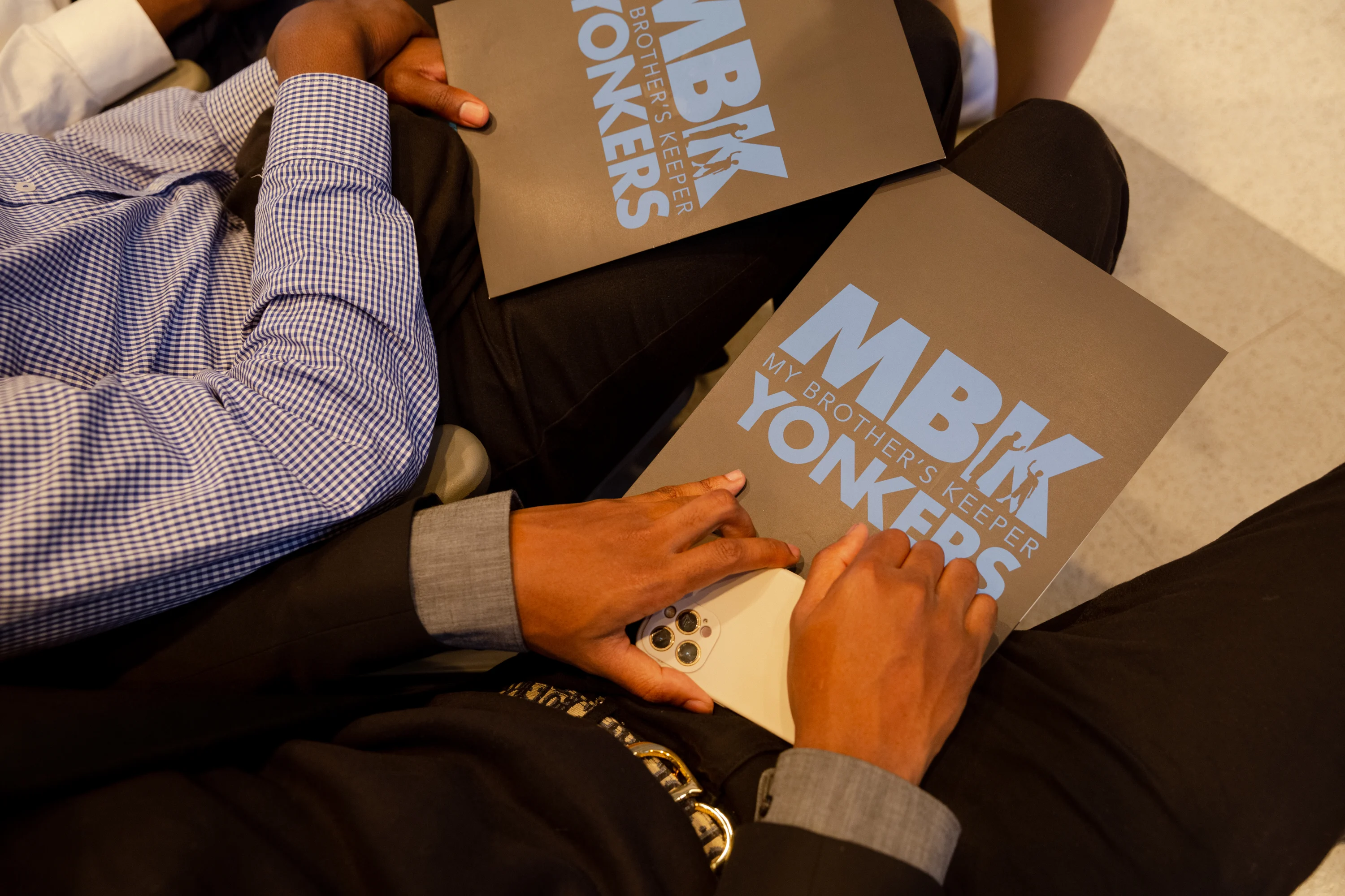 An image of two brown hands with a deep skin tone hold folders that read, “MBK Yonkers” and “My Brother’s Keeper.” 