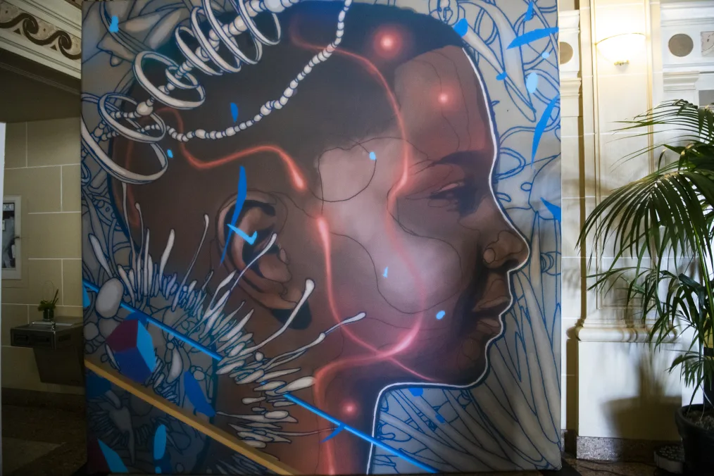 A photo of a piece of art work off to the side of a well decorate floor of an office building. The art is a side profile of a young woman whos hair at the top is short and dark brown. The sides of her hair fade and get lighter in color. She has a deep skin tone and is lightly smiling. She is surrounded by  silver, iron, grey, and white painted rainforest elemnents and patterns.