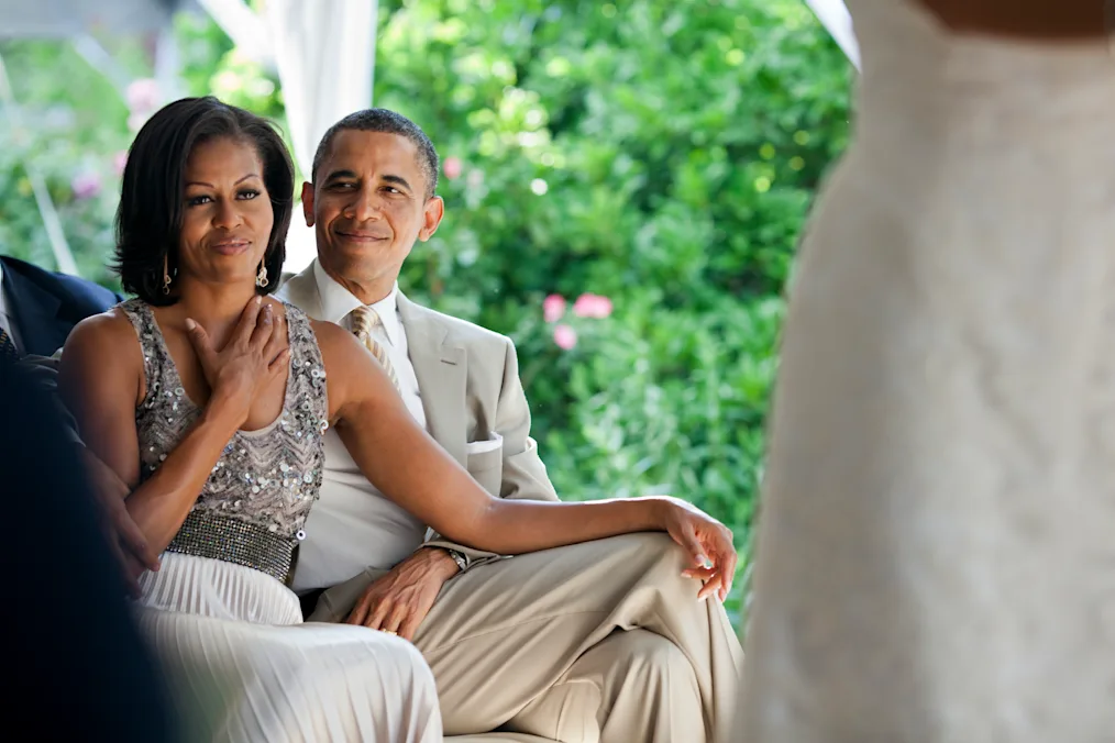 President Obama, in a tan suit, smiles at his wife, who is seated next to him. Former First Lady Michelle Obama grabs her husband’s knee with her left hand and rests her right hand on her neck.