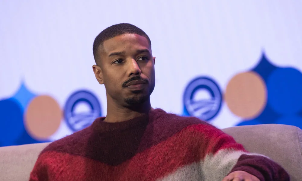 Micheal B. Jordan, a black man with a medium deep skin tone, mustache, and low hair cut wearing a red, maroon, and white striped sweater looks to the right. T