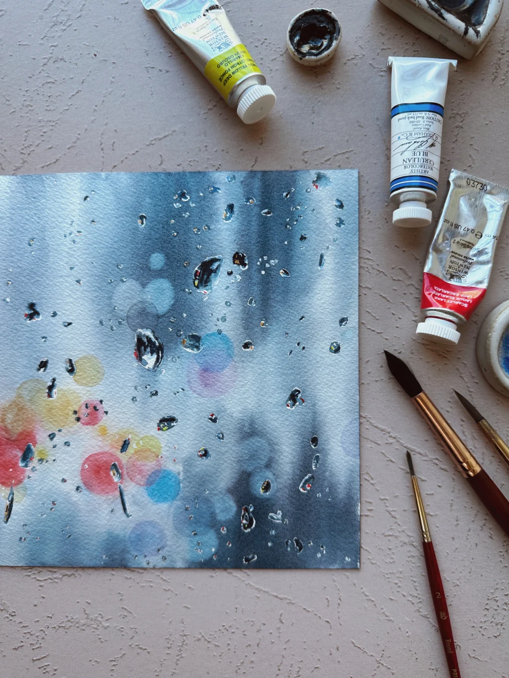 An image of a watercolor painting created by Kolbie Blume. The picture is blue and white with dots of red, pink, yellow, and blue. The picture also has painted raindrops on it. Next to the painting are to paint tubes, and three paint brushes.