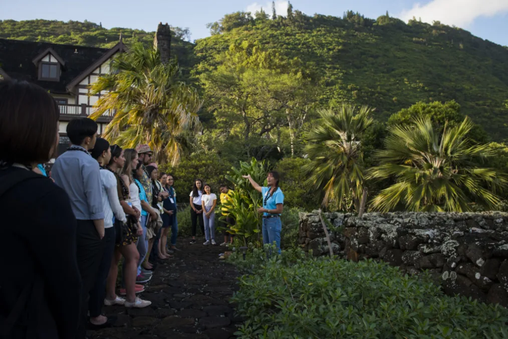 A group of people with a range of light and light medium skin tones stand outdoors and listen to a guide in a bright blue shirt and jeans.