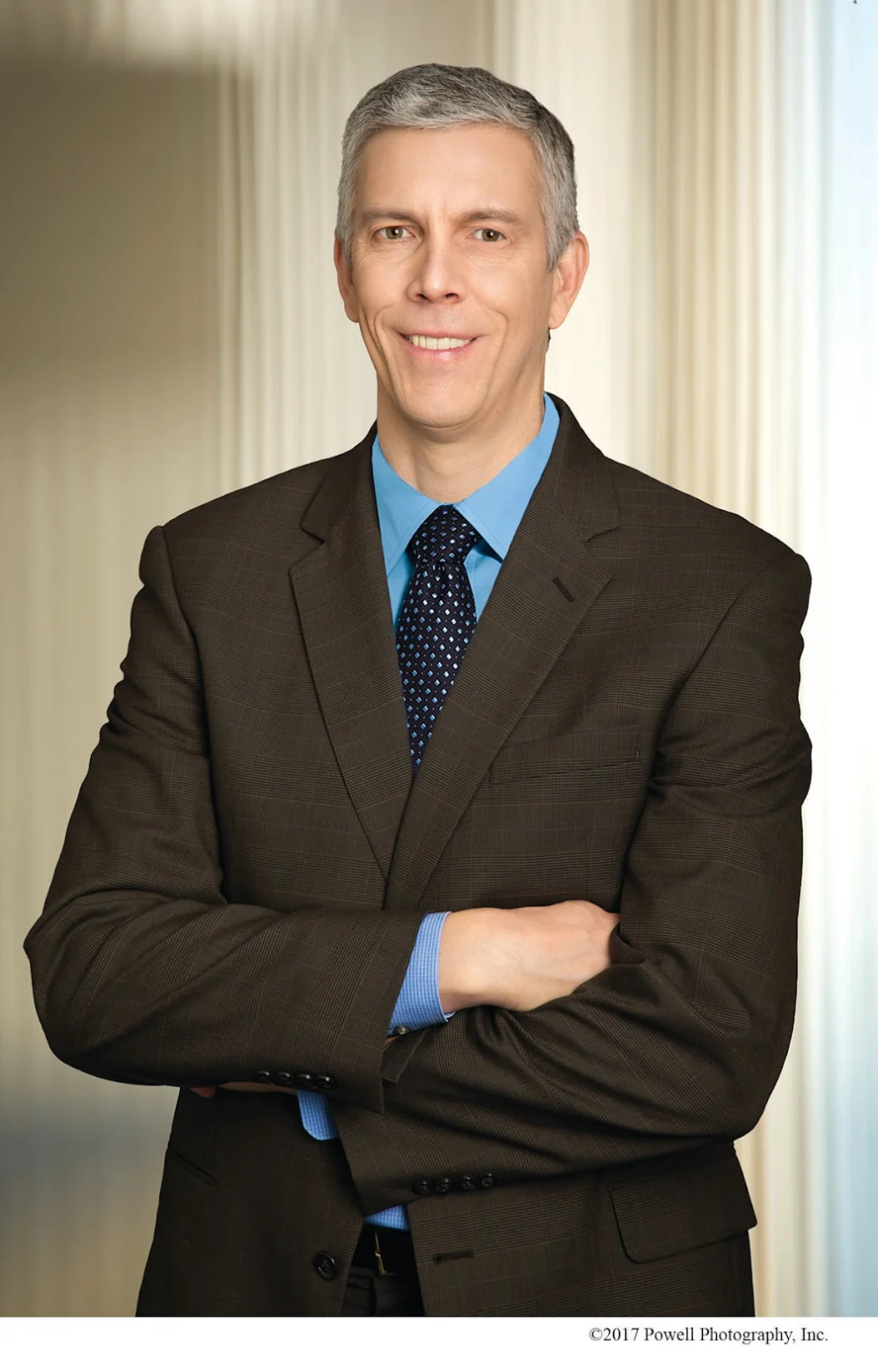 A waist-up headshot of a White man with gray hair, blue shirt and tie and dark suit jacket. His arms are crossed and he smiles slightly, showing teeth.