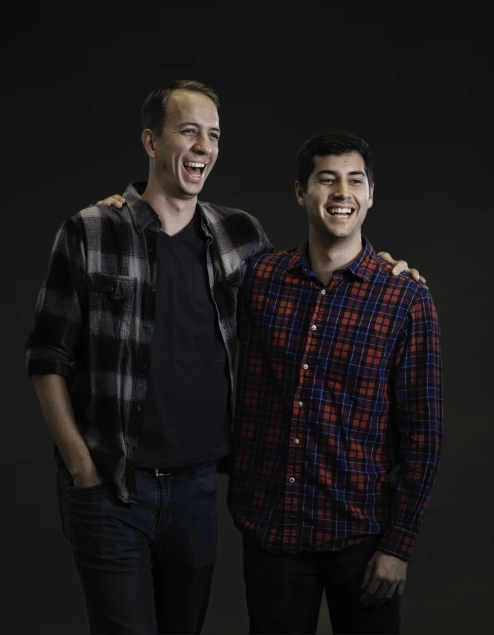 Two men with light skin tones laugh and smile while wearing plaid dress shirts and jeans.