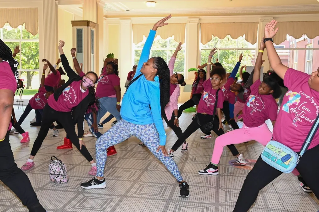 A Black woman wearing a blue sweatshirt and floral leggings holds a yoga pose. She is encircled by a group of adolescent Black girls wearing matching maroon shirts and holding the same pose. They are a range of light to dark skin tones. 