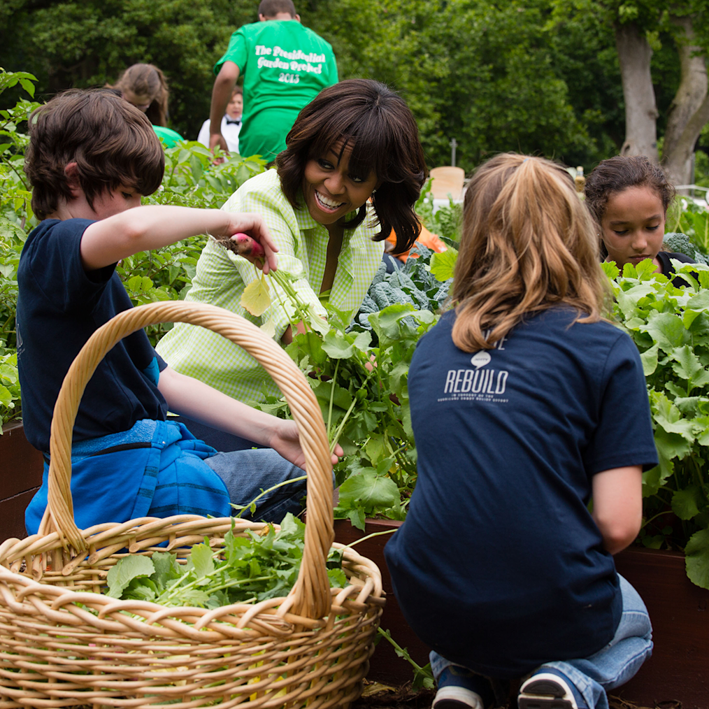 Mrs. Obama wears a green top as she gardens with two young people at the White House Garden. The young boys and the girl have light and dark tones and brown hair. 