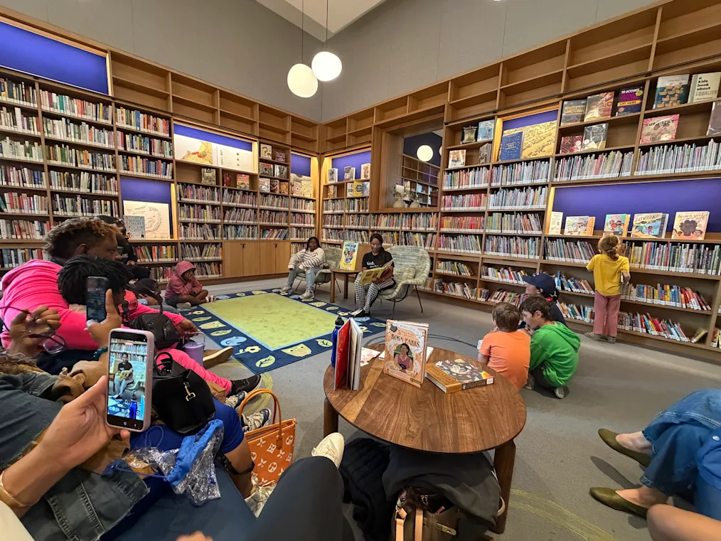 Ebony Wilkins, A Black woman with a dark skin tone and box braids, sits as she reads to a group of children with a range of light to dark skin tones at the Chicago Public Library Branch at the Obama Presidential Center.