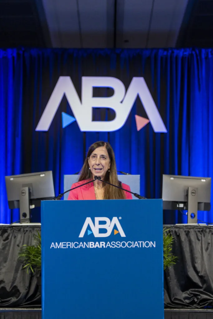 Mary Smith speaks from behind a blue podium. The words on the podium read “American Bar Association” along with its logo. In the background, there are computer monitors and royal blue drapes.