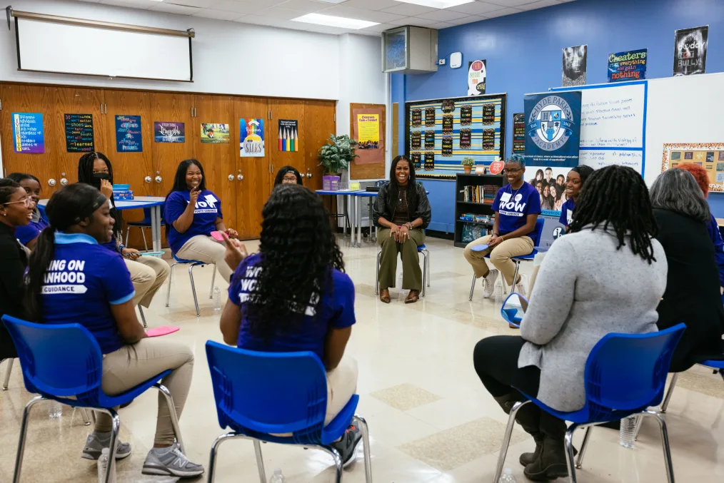 Michelle Obama leads a circle group with a mix of young and older women of deep skin tones. Michelle Obama is wearing a green and brown themed business causal attire. The women in the group are wearing blue "Wow youth guidance" shirts and kahkis. Some of the girls, including Michelle Obama, spot the camera and start smiling and or pointing.