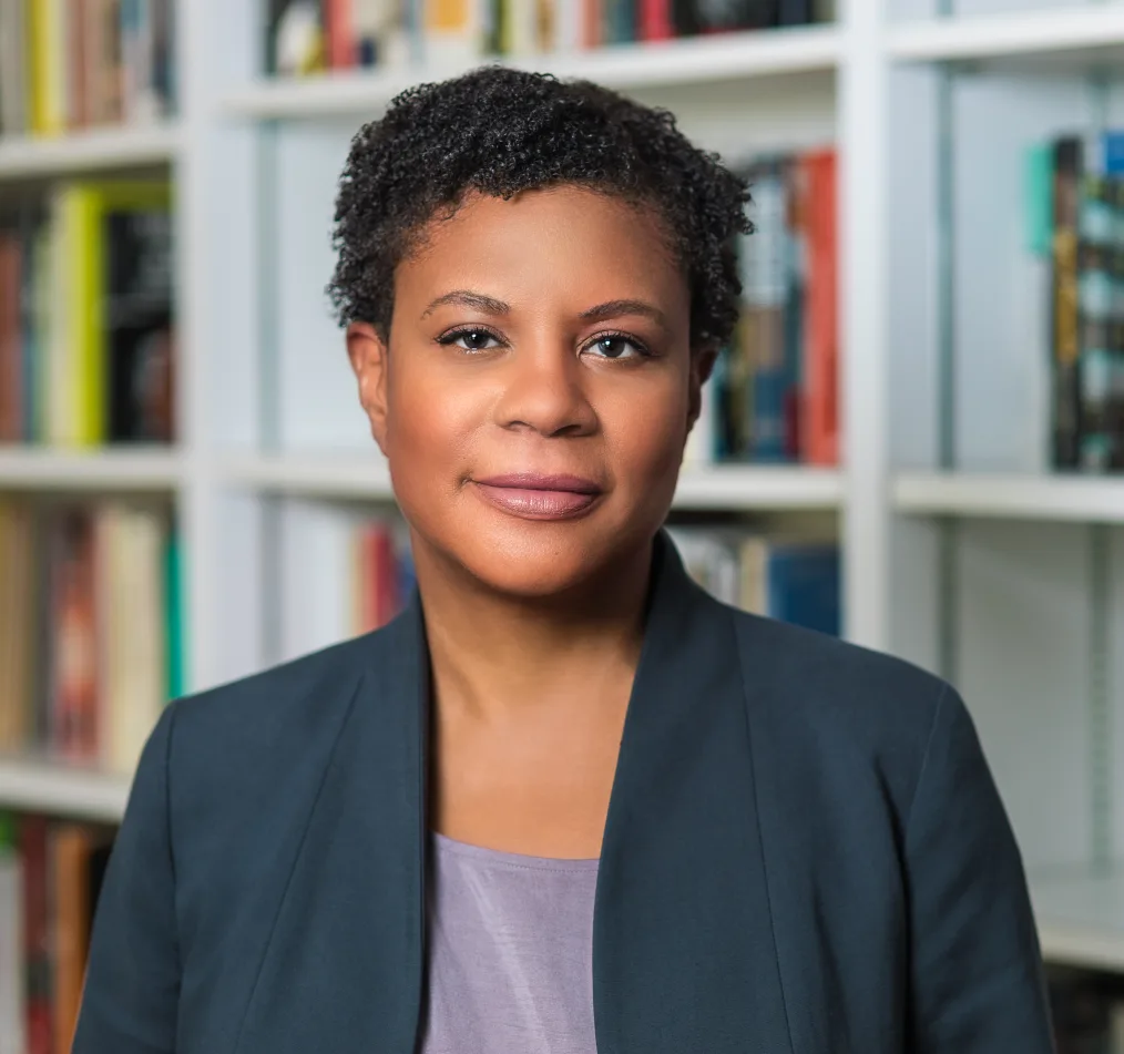 A Black woman with short, curly black hair smiles slightly, without showing her teeth. She wears a dark gray suit jacket over a purple top. Behind her are white bookshelves with colorful books.