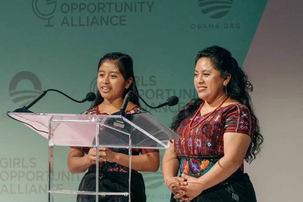 Maria Florinda Meletz Chumil and Vilma Saloj stand behind a clear podium and speak at the Girls Opportunity Alliance Get Her There campaign event. Both have medium skin tones, dark hair, and are wearing traditional Guatemalan clothing, rich with jewel-toned colors and intricate patterns.