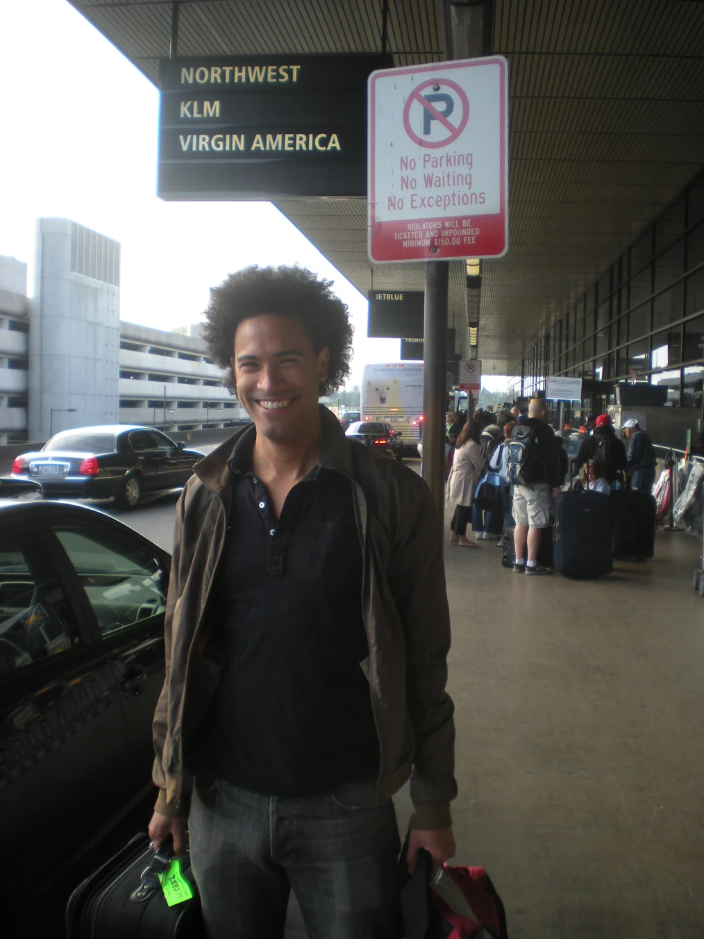 Devin Hampton, a Black man with a light medium skin tone, smiles outside of an airport terminal. In this photo, he has a short afro and is holding a bag, wearing a black shirt and cargo jacket. 