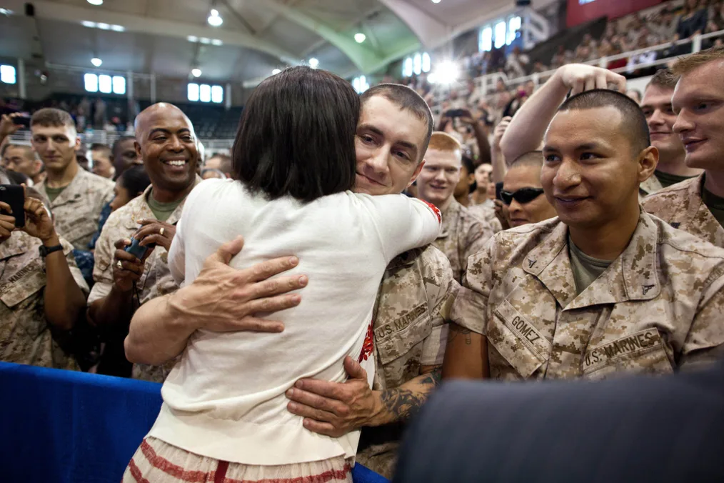 A woman with a white and red sweater hugs a man with a light skin tone wearing a military uniform. There is a large group of other men with a variety of skin tones around the, also wearing military uniforms.  