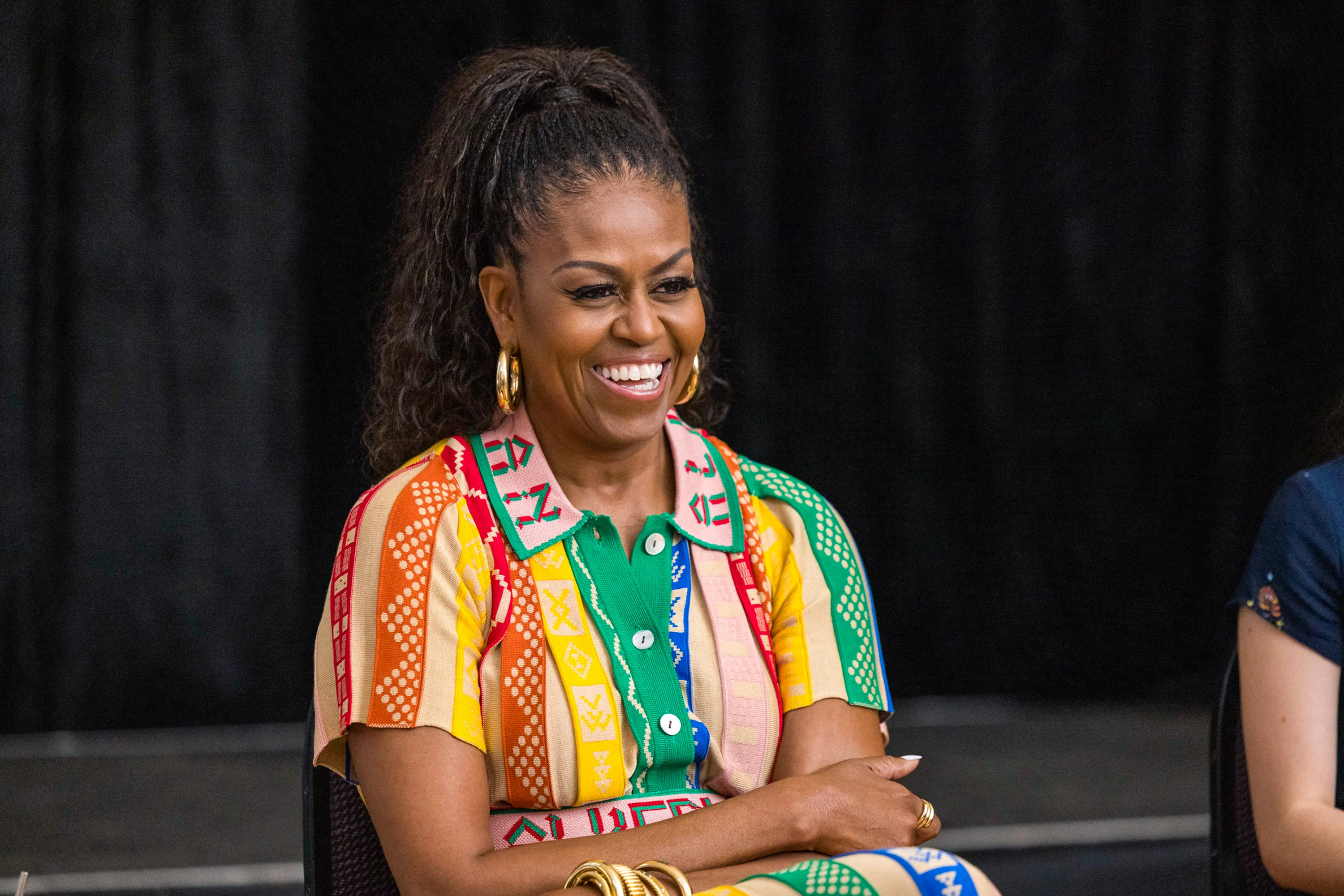 Michelle Obama laughs. She is wearing a colorful dress and has her arms crossed.