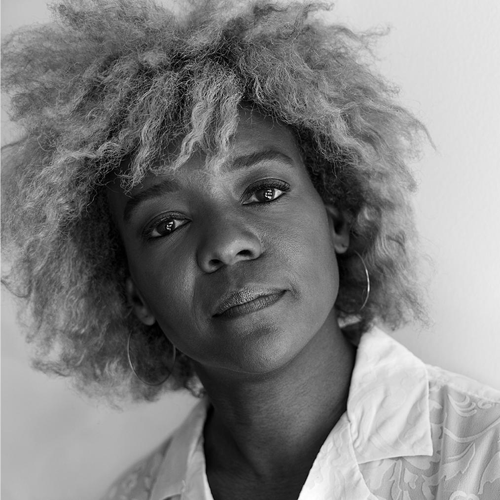 A photographed portrait of Nekisha Durrett. The image is in black, white, and greyscale tones. Nekisha has a deep complexion and light colored hair that falls in waves around the nape of her neck. She is wearing hoop earrings and a white collared shirt with an embroidered design.