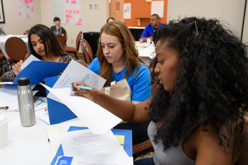 In this picture, multiple people with various skin tones are in a classroom looking space with pens
and paper looking like they're participating in some sort of project.