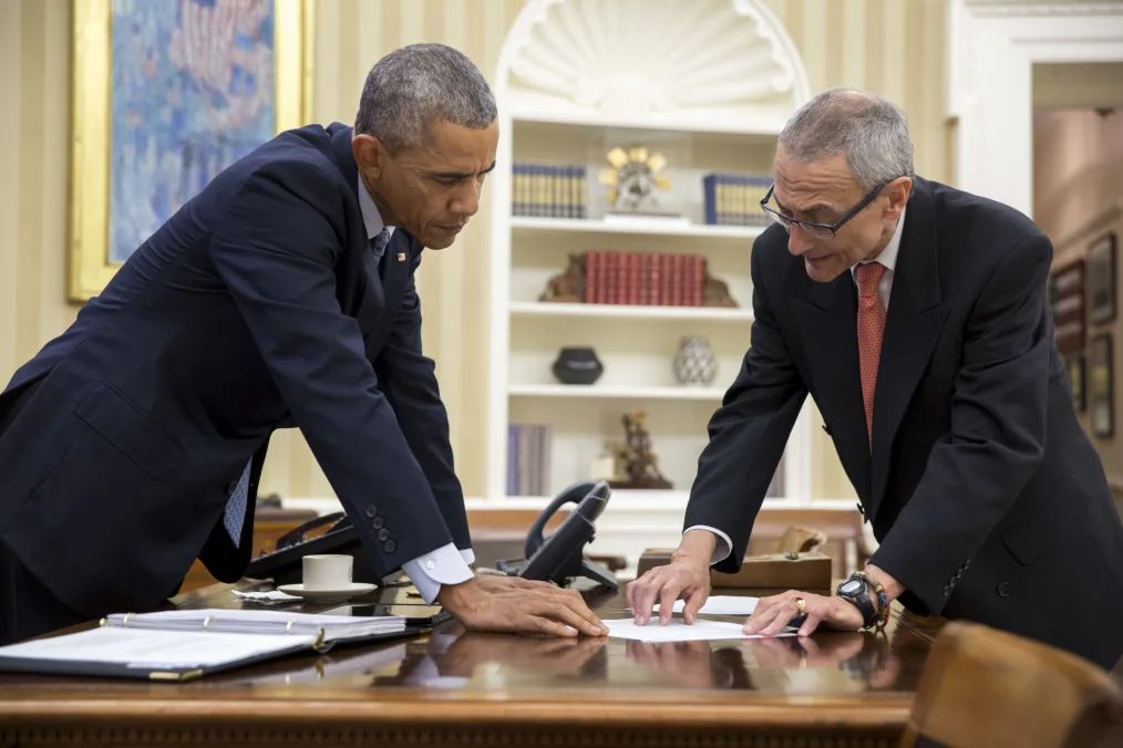 President Obama leans over his desk with a concentrated face at a document while an older man with a light skin tone , gray short hair and glasses looks with him in business attire 